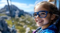 Young girl wearing blue glasses smiles joyfully while riding cable car in mountains, wind blowing through her hair against backdrop of scenic alpine peaks and summer sky.