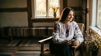 Young woman in traditional embroidered white blouse sitting by window in rustic interior, warm natural lighting creating cozy atmosphere for lifestyle content.