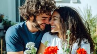 Happy mixed-race couple sharing intimate moment with flowers in modern home interior. Young man and woman embracing, laughing together in bright living space.