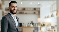 Confident male professional with dark hair and beard wearing gray business suit and white shirt, smiling at camera in modern office kitchen environment with blurred background.