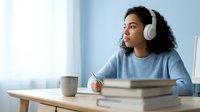 Young woman with curly dark hair and medium skin tone wearing wireless headphones and light blue sweater, studying at wooden desk with books and coffee cup, suitable for education and learning content.