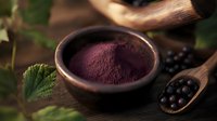 Ground acai berry powder in wooden bowl with fresh berries on spoon, dark moody food photography on rustic wooden surface with green leaves.