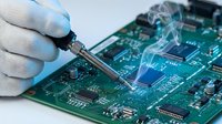 Technician in white gloves soldering a circuit board with rising smoke, close-up of electronics repair and manufacturing process.