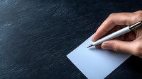 Female hand holding silver pen ready to write on blank white paper against dark textured background, copy space for business or education concepts.