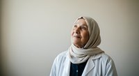 Senior woman wearing beige headscarf and light blue jacket, looking upward with contemplative expression against neutral background for healthcare, wellness, and lifestyle content.