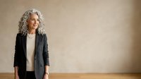 Mature woman with gray curly hair wearing black blazer and white top standing against neutral beige wall background, professional portrait for business, corporate, and lifestyle design projects.
