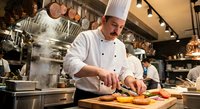 Professional chef in white uniform and hat preparing fresh ingredients on wooden cutting board in busy commercial restaurant kitchen with steam and copper cookware.