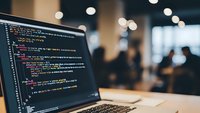 Laptop displaying colorful code on screen in modern office workspace with blurred background of colleagues working in contemporary business environment.