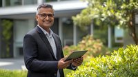 Confident middle-aged South Asian businessman in suit holding digital tablet outdoors, smiling against modern office building with green landscaping.