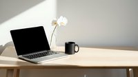 Laptop with blank screen on wooden desk next to white flower in vase and black mug, minimalist workspace setup for business and design projects.