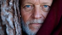 Close-up portrait of mature Caucasian man with intense blue eyes and gray beard looking through wooden texture, dramatic lighting creates mysterious atmosphere.