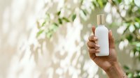 Unisex hand holding white cosmetic bottle with gold cap against blurred green foliage background, suitable for skincare, beauty, wellness product marketing.