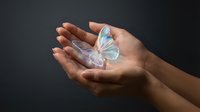 Cupped hands gently holding luminous holographic butterfly with iridescent wings against dark background symbolizing hope, transformation and spiritual growth.