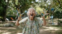 Joyful senior woman with paint-covered hands laughing outdoors in garden setting, showcasing creativity and vibrant spirit in retirement.