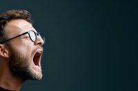 Side profile of young Caucasian man with glasses and beard yawning or screaming against dark background, expressing tiredness, stress or strong emotion.