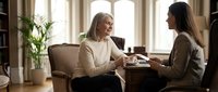 Two women having a meaningful conversation over coffee in an elegant living room, senior Caucasian woman and young Asian professional engaged in warm discussion.