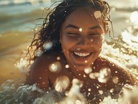 Young African American woman laughing with joy while swimming in ocean at sunset, water droplets and golden bokeh light creating magical summer atmosphere.
