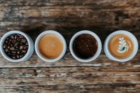 Four white cups on rustic wood showing coffee stages: roasted beans, espresso, ground coffee, and latte art. Top view cafe concept.