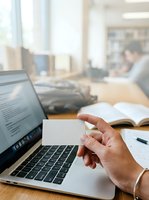 Caucasian male professional holding blank business card at wooden desk with laptop, open notebook, and colleagues working in blurred office background for corporate branding.