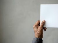 Mature male hand holding blank white card against neutral gray wall background for messaging or advertising.