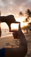 Hands forming frame gesture at tropical beach sunset with palm trees silhouettes, creative photography composition for travel and vacation concepts.