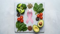 Pink ribbon surrounded by healthy foods including broccoli, spinach, avocado, tomatoes, seeds and nuts on white marble background for cancer awareness.