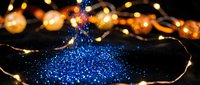 Blue glitter sparkles in dark bowl with fairy lights creating magical bokeh background for holiday decorations and festive celebrations.
