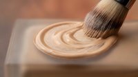 Paintbrush applying swirled beige stain or wood finish on light surface, showing application technique for home improvement and DIY projects.
