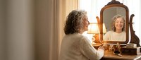 Mature Caucasian woman with curly gray hair smiling at reflection in vintage wooden vanity mirror, enjoying positive self-care moment in warm bedroom.