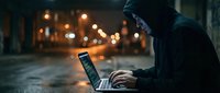 Hooded figure using laptop computer on dark city street at night with blurred lights background for cybersecurity and hacking concepts.