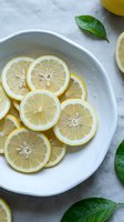 Fresh lemon slices arranged in white ceramic bowl with green basil leaves on light textured surface for healthy cooking and natural wellness concepts.