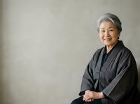 Senior Asian woman with gray hair wearing dark textured jacket, sitting against neutral background, smiling at camera. Portrait suitable for healthcare, wellness, lifestyle, and corporate communications.