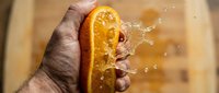 Male hand squeezing fresh orange half with juice splashing, isolated on blurred background. Suitable for food, beverage, and healthy lifestyle content.