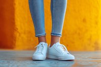 Female legs in blue skinny jeans and white casual sneakers standing against vibrant orange wall background, modern urban street style fashion photography.