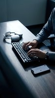 Male professional with dark skin tone typing on keyboard at desk with headphones and smartphone, suitable for business, technology, and work concepts.