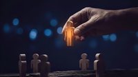 Hand selecting glowing orange human figure from wooden people icons on dark background with bokeh lights for recruitment and leadership concepts.