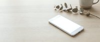 White smartphone on wooden desk with eucalyptus branch and coffee cup in minimalist workspace setup for modern business and technology concepts.