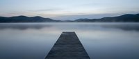 Wooden dock extending into calm misty lake at dawn with mountain silhouettes creating peaceful meditation and relaxation atmosphere for wellness concepts.