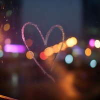 Heart drawn on foggy window glass with colorful bokeh city lights in background creating romantic atmosphere for Valentine's Day celebrations.