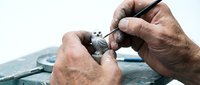 Skilled craftsman hands engraving intricate details on silver jewelry piece using precision tools at workbench for custom metalwork and artisan craftsmanship.