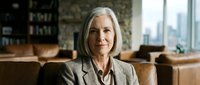 Professional mature Caucasian businesswoman with gray hair in elegant suit sitting confidently in modern office with city skyline view and leather furniture.