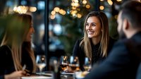 Young woman with bright smile enjoying dinner with friends at upscale restaurant, warm ambient lighting creates intimate atmosphere for social gathering.