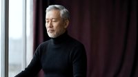 Mature Asian man with gray hair in black turtleneck sweater sitting peacefully by window with eyes closed, meditation and mindfulness concept for wellness.