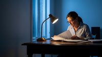 Young professional woman working late at office desk with lamp in blue evening light reading documents and paperwork for business project deadline.