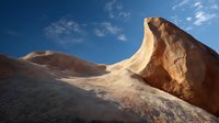 Dramatic rock formation in desert landscape with smooth curved surfaces against vibrant blue sky with wispy clouds.