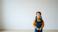 Young Asian girl with braided hair wearing yellow long-sleeve shirt and denim overalls standing against white wall, suitable for education and childhood lifestyle content.