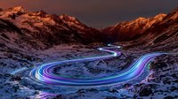 Long exposure light trails curve through snowy mountain valley at dusk, creating glowing blue and purple streaks across winding road below dramatic peaks.