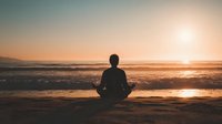 Silhouette of person meditating on beach at sunset, sitting in lotus position with ocean waves and golden light creating peaceful atmosphere.