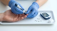 Healthcare professional in blue gloves performing blood glucose test with lancet device on patient hand, diabetes monitoring equipment on medical tray.