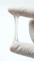 Close-up of clear gel or serum stretching between two fingers wearing white gloves, demonstrating viscosity and elasticity for skincare, cosmetics, or laboratory testing purposes.